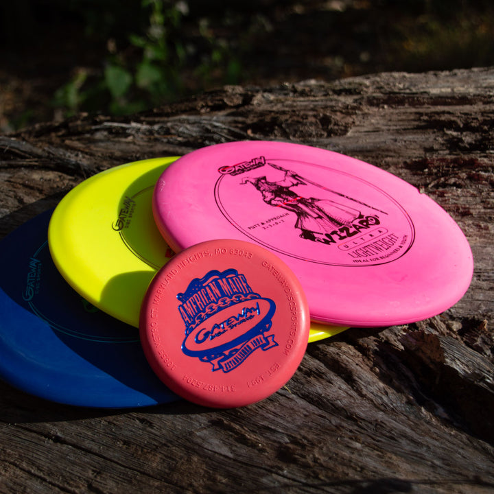 The 3-Disc Ultra Lightweight Starter Set with Mini, featuring pink, yellow, blue, and red discs including a putter, is stacked on weathered wood outdoors in sunlight with soft shadows.
