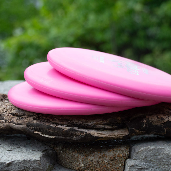 Three pink plastic Voodoo - Money discs are stacked on a piece of wood, with a blurred green background.