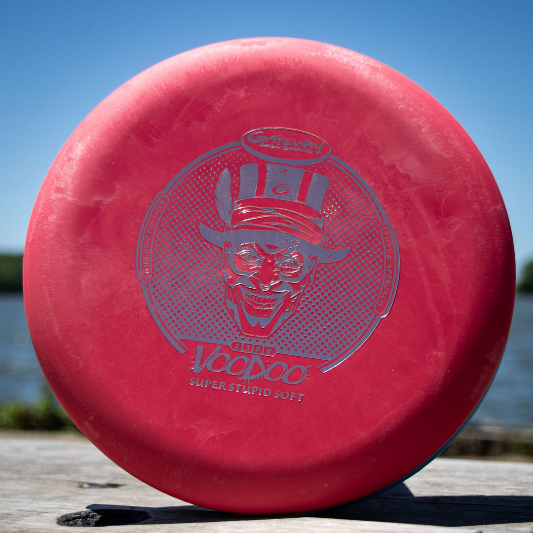 A red Voodoo - Suregrip - SSS putter with a grinning skeleton in a top hat graphic, displayed on wood beside water.
