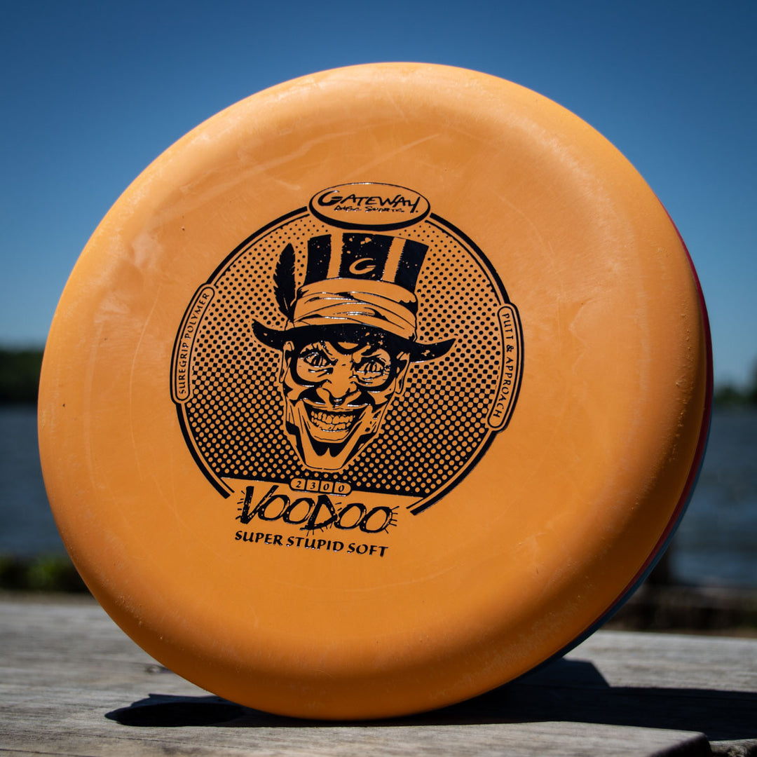 An orange Voodoo - Suregrip - SSS disc golf putter stands upright on wood with water and trees in the background—ideal for straight-line putts or reliable lay-ups.