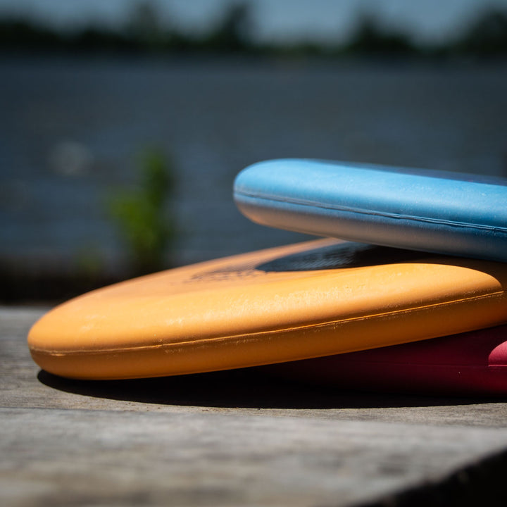 Close-up of stacked Voodoo - Suregrip - SSS orange and blue lay-up discs on a wooden surface, blurred water in the background.