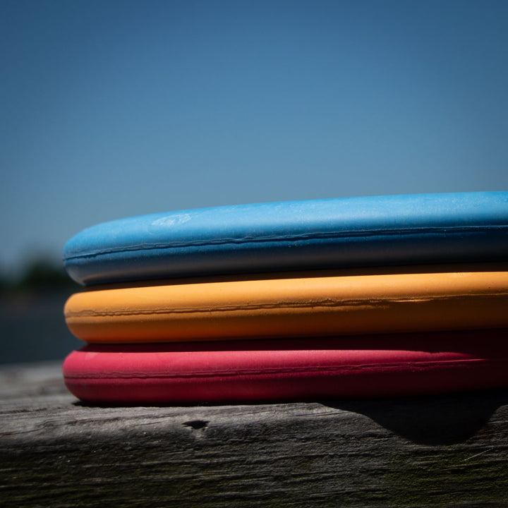 Three Voodoo - Suregrip - SSS foam kickboards in blue, yellow, and red are stacked on a wooden surface with a blurred outdoor background, evoking the classic color scheme of vintage sports gear.