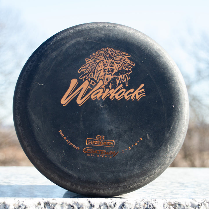 A black Warlock - Suregrip - SS putter with gold logo and text stands upright on a light-colored outdoor surface.