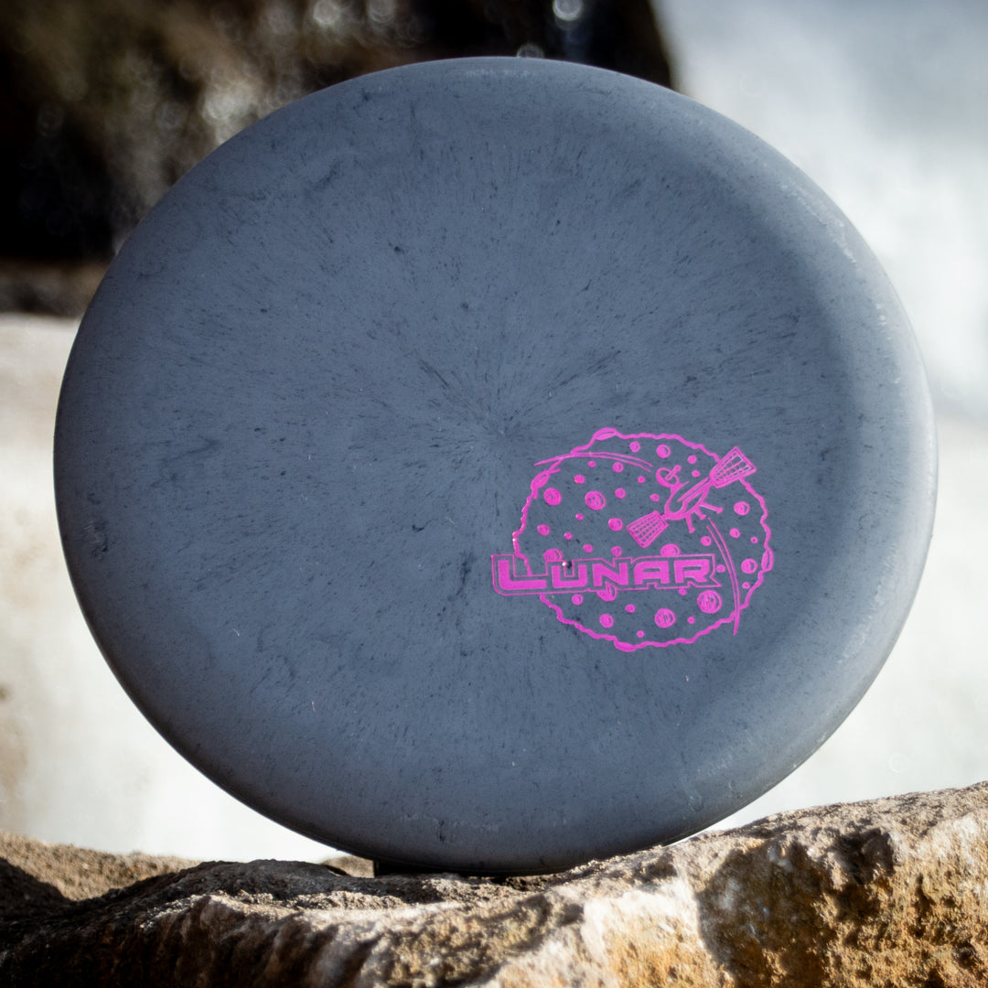 A gray Warlock - Lunar disc golf putter with a pink "Lunar" logo and lunar graphic rests on a rock outdoors.