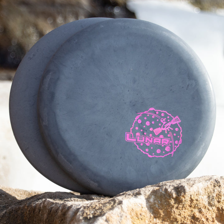 Two dark gray Warlock - Lunar disc golf putters lean against a rock; one displays a pink "LUNAR" graphic with a moon illustration, evoking the style of the renowned Warlock and Wizard series.