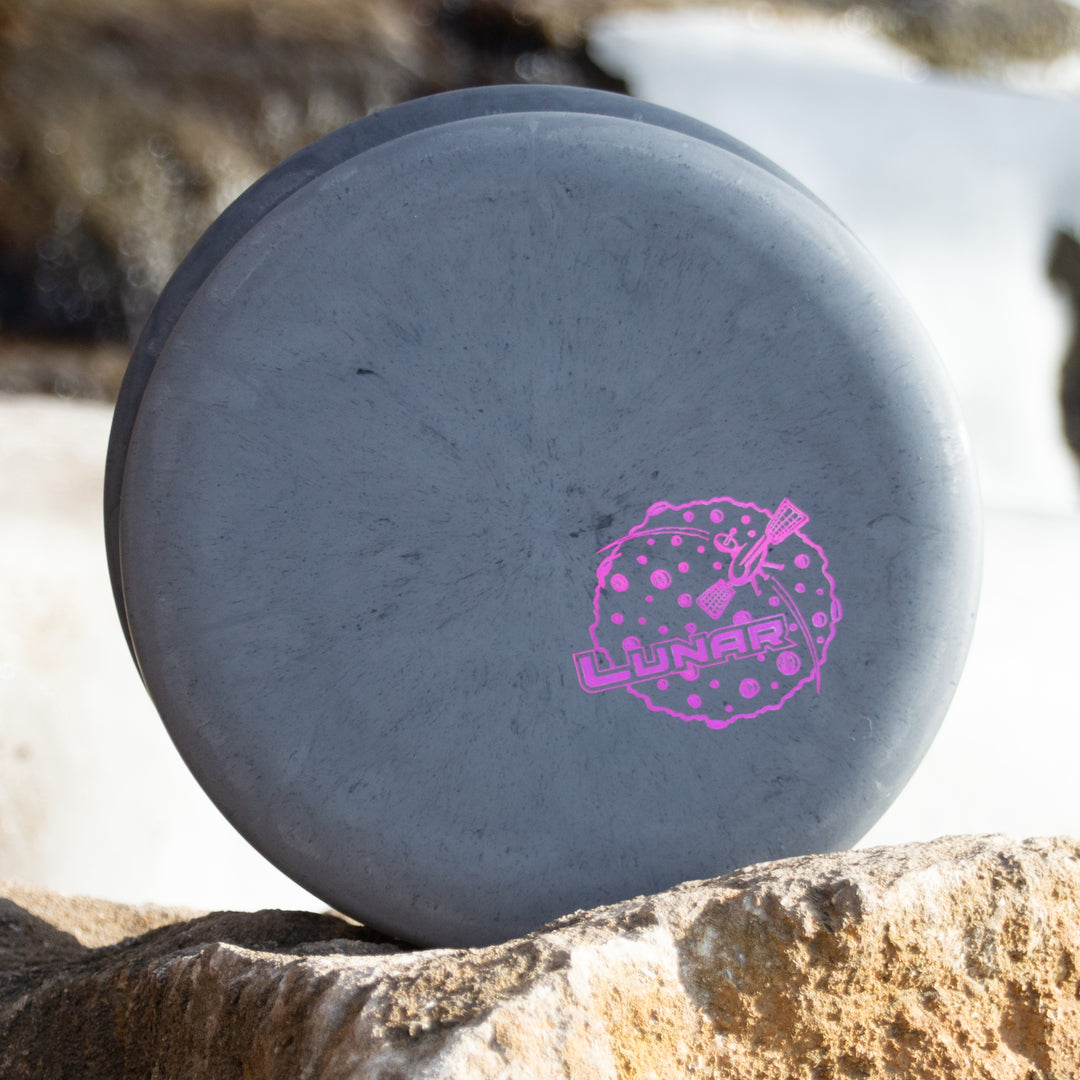 The Magic - Lunar is a gray disc golf putter with "Lunar" in pink, famous for its straight and gliding flight, pictured resting on a rock in front of a blurred outdoor background.