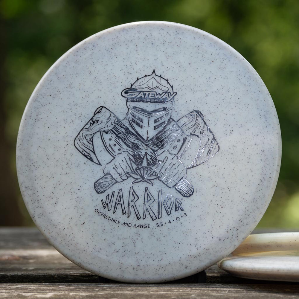 A white Warrior - Diamond Hemp mid-range disc with an armored warrior sketch and "Gateway Discs Warrior, Overstable Mid-Range, 5 4 0 3" text rests on a wooden surface outdoors, showcasing its overstable flight and consistent glide.
