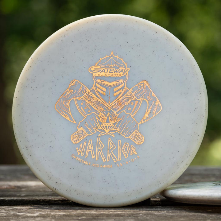 A white Warrior - Diamond Hemp overstable disc golf disc featuring a gold warrior with two axes and "Gateway Disc Sports Warrior, Overstable Mid Range" text rests on a wooden surface.