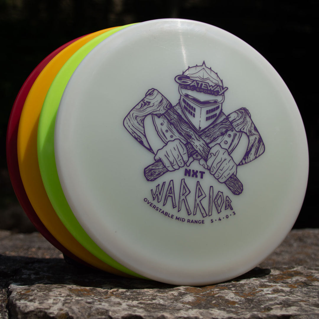 Four stacked disc golf discs rest on a rock, with the front Warrior - NXT mid-range disc displaying a knight illustration and the text "Warrior - NXT Overstable Mid Range 5 4 0 3.