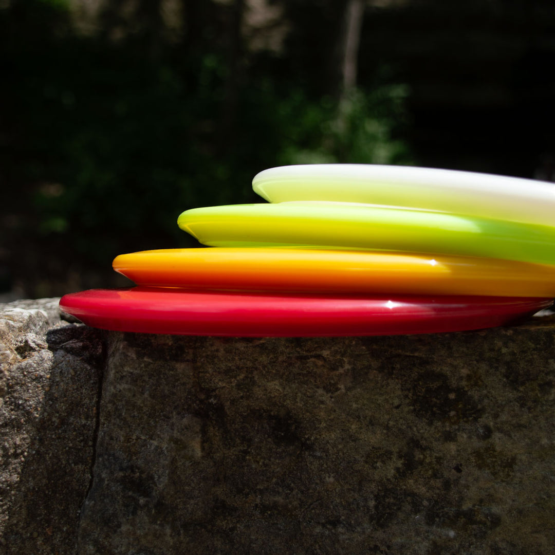 Four vibrant frisbees—red, orange, yellow, and white—are stacked outdoors on stone with greenery behind them, featuring the Warrior - NXT mid-range disc famous for its over-stable flight.