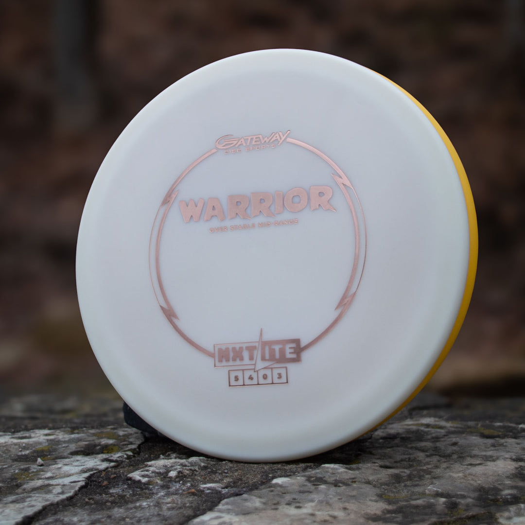 The Warrior - NXT Lite, a white and yellow mid-range disc golf disc by Gateway, stands upright on a rock, ideal for hyzer approaches against a blurred natural background.
