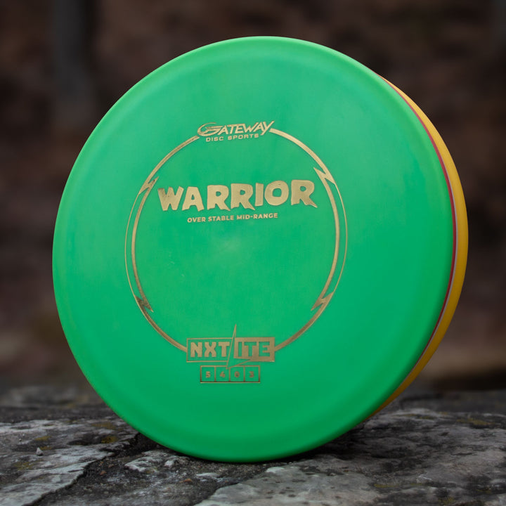 A green Gateway Warrior - NXT Lite mid-range disc with gold lettering rests on a stone surface, ideal for hyzer approaches.