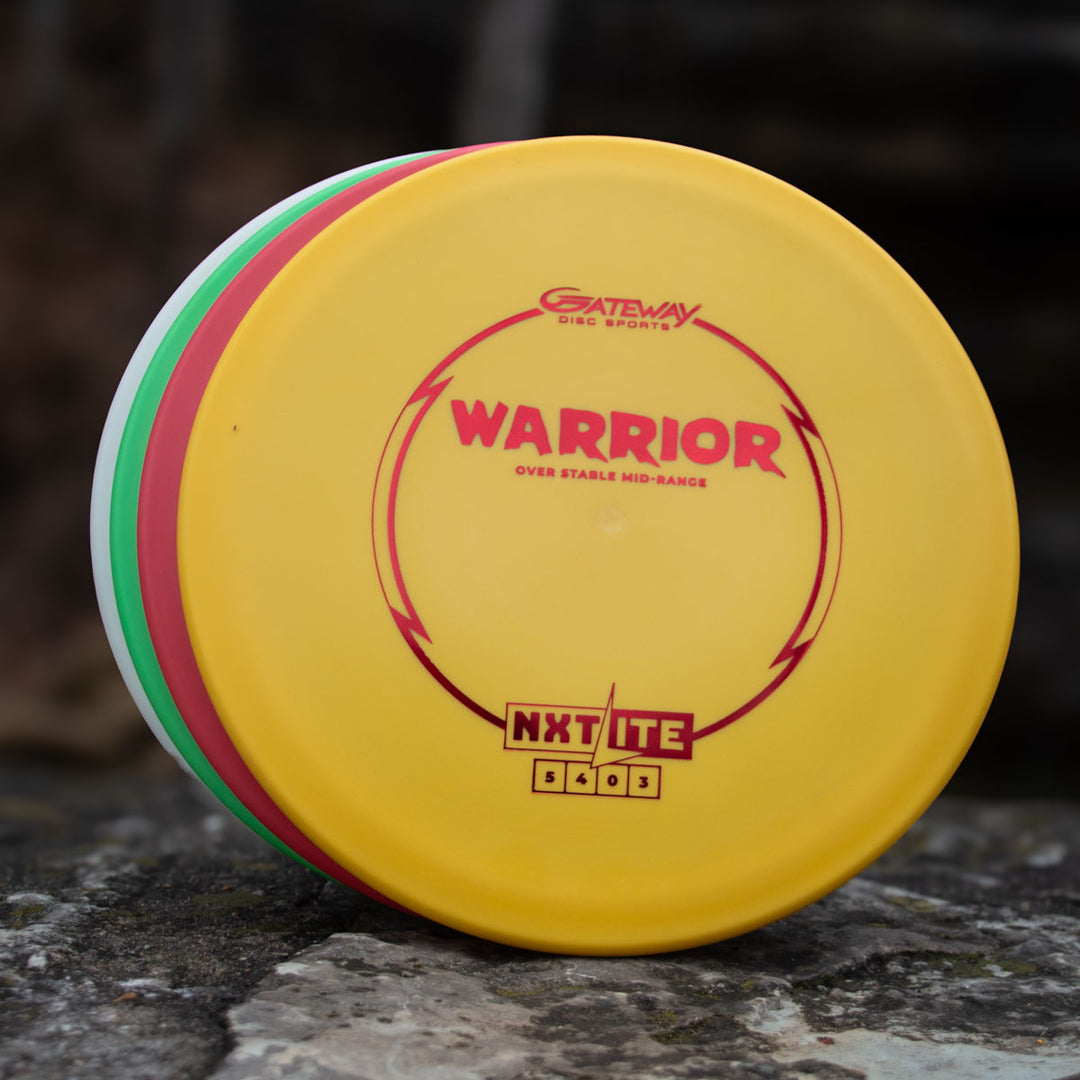 Three disc golf discs sit stacked on a rock; the top yellow disc, labeled "Warrior - NXT Lite" by Gateway Disc Sports, is a dependable mid-range ideal for controlled hyzer approach shots.
