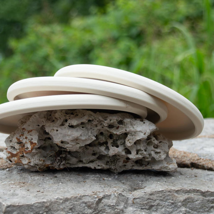 Three Warspear - Suregrip white ceramic plates with a low profile are stacked on a porous rock outdoors, with greenery in the background.