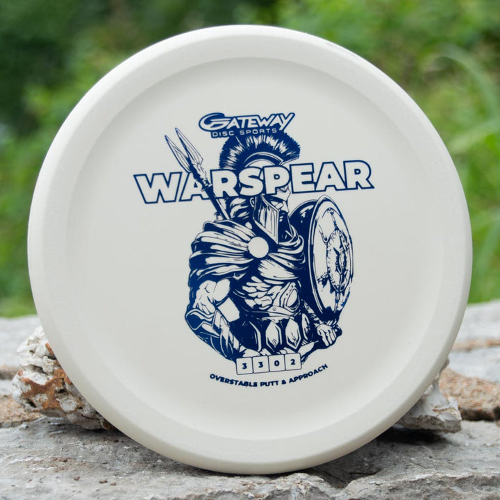 The Warspear - Suregrip is a white, low profile putt and approach disc featuring a Spartan warrior holding a shield, standing on a rock surrounded by greenery.