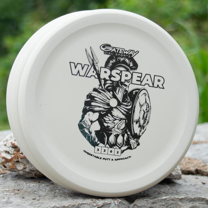 A white, low-profile Warspear - Suregrip disc golf putter by Gateway Disc Sports, featuring a Spartan warrior design, rests on a rock—perfect for dependable putt and approach shots.