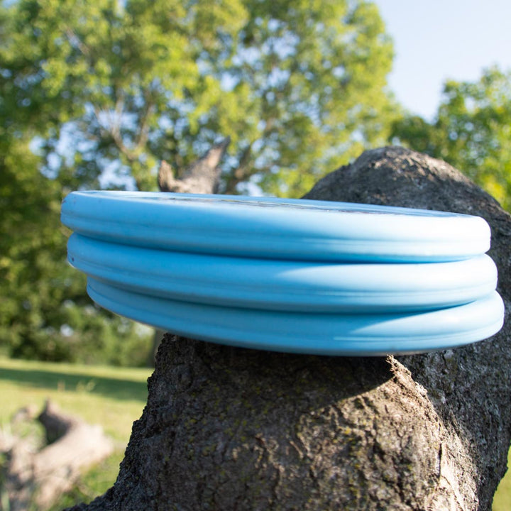 Three light blue Warspear - Suregrip Nylon discs with Thumb Track design are stacked on a tree branch, set against lush green trees and a bright blue sky.
