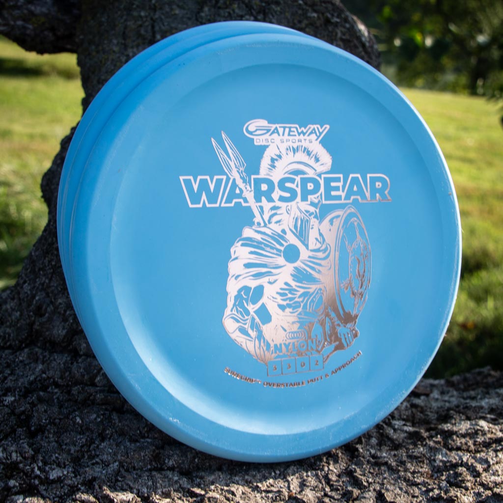 A blue Warspear - Suregrip Nylon putt and approach disc with a silver illustration and Thumb Track design rests against a tree trunk outdoors on a sunny day.