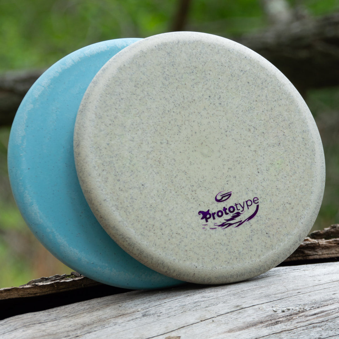 Two Witch Doctor Proto discs, light blue and light gray with "Prototype 44x" in purple, rest on wood outdoors—ideal for fans of beaded throwing putters.