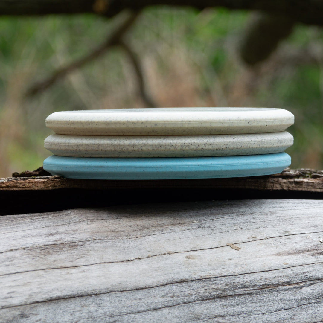 Three round, flat Witch Doctor Proto soap bars—two off-white and one light blue—are stacked on weathered wood, their smooth surfaces like a beaded throwing putter, set against a blurred outdoor background.