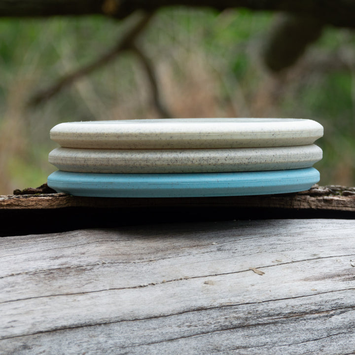 Three round, flat Witch Doctor Proto soap bars—two off-white and one light blue—are stacked on weathered wood, their smooth surfaces like a beaded throwing putter, set against a blurred outdoor background.