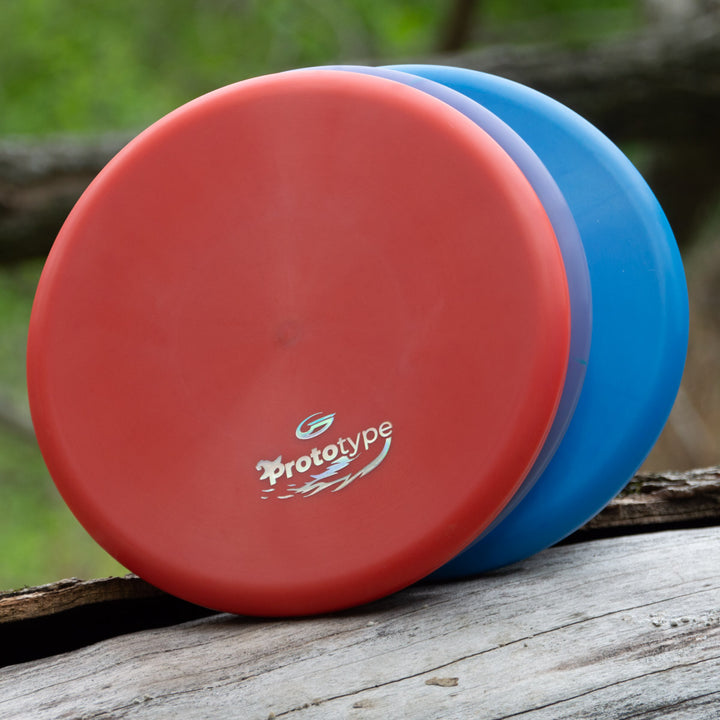 A red Witch Doctor Proto disc labeled "Prototype 44x" is stacked on a blue disc outdoors on a weathered wooden surface, resembling a beaded throwing putter.