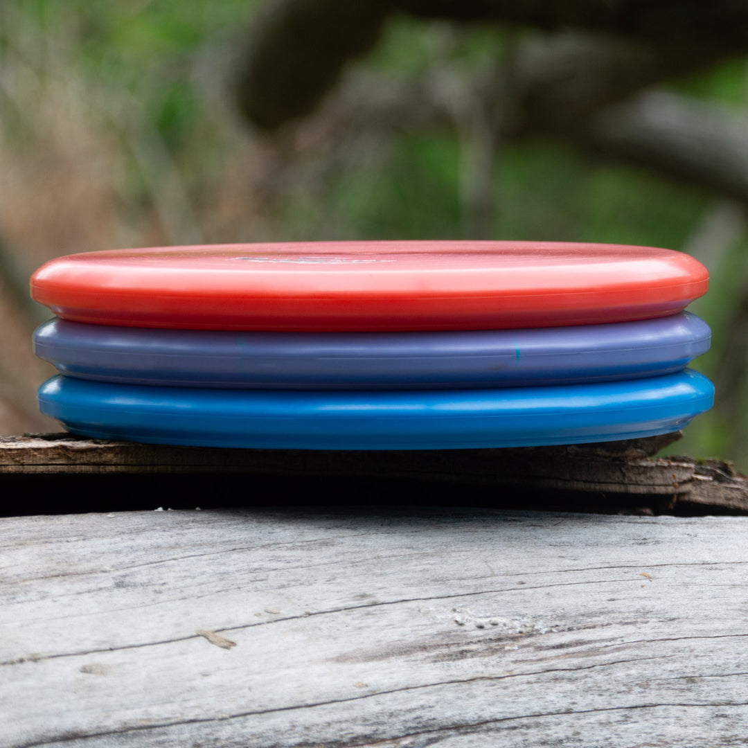 Three disc golf discs—red, purple, and blue—stacked on a weathered wooden surface outdoors, featuring the Witch Doctor Proto beaded throwing putter.