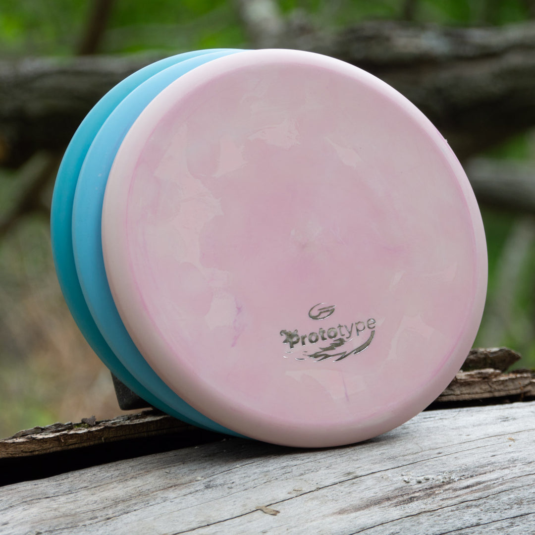 Two disc golf discs, one pink and one blue, rest on a weathered log outdoors. The pink disc is a beaded throwing putter from the Witch Doctor Proto line.