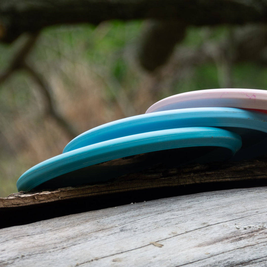 Three flying discs—two blue, one pink—are stacked on a fallen log outdoors with blurred trees behind. The top disc is the Witch Doctor Proto, a beaded throwing putter.