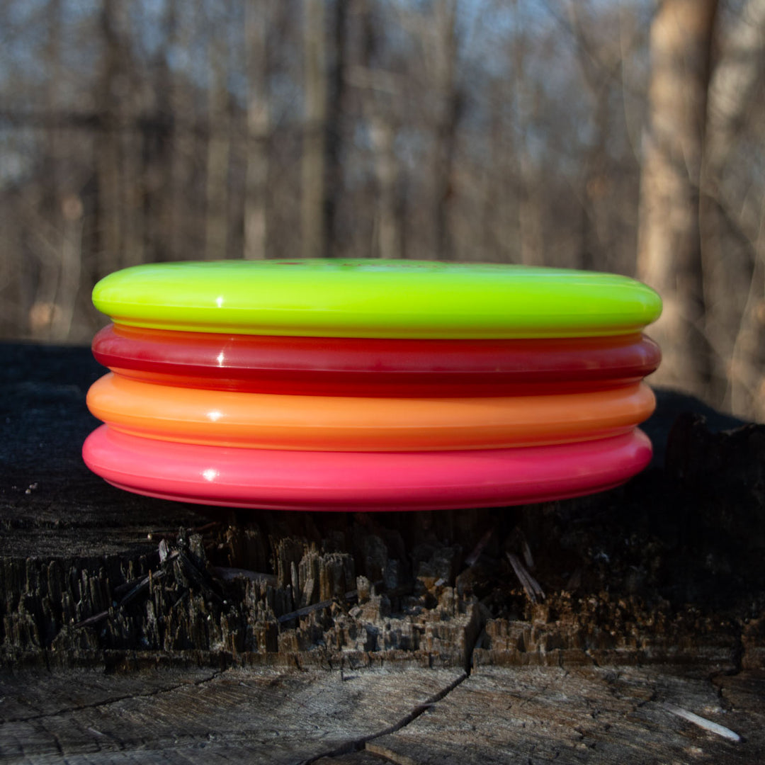 Four Wizard - Evolution - Neon disc golf discs in vibrant Day Glow Colors—yellow, orange, and pink—are stacked on a tree stump outdoors with a blurred forest background.