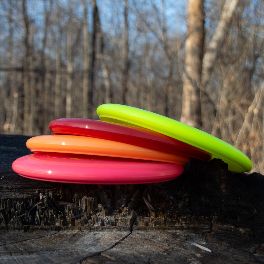 Four brightly colored Wizard - Evolution - Neon disc golf discs are stacked on a tree stump in the woods. The Day Glow Colors make them beautifully stand out against the bare trees and natural setting.