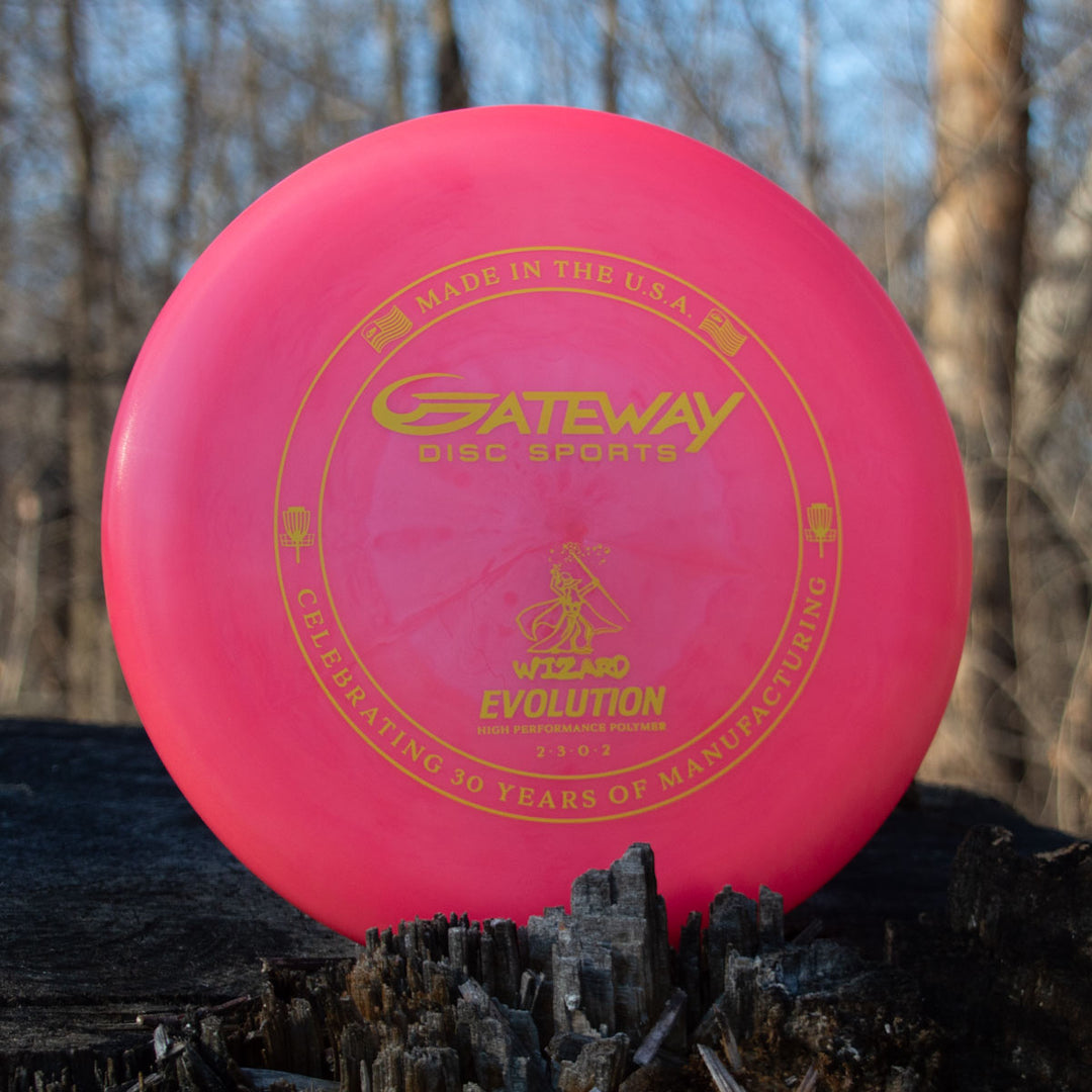 A pink Wizard - Evolution - Neon disc by Gateway Disc Sports with gold text and graphics stands upright on wood outdoors, showcasing Day Glow Colors with trees in the background.