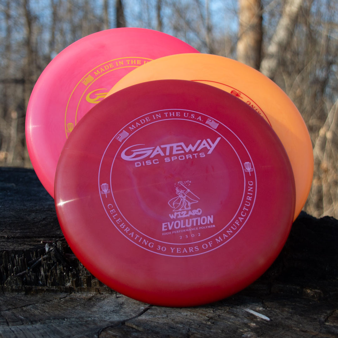 Three vibrant Gateway Disc Sports Wizard - Evolution - Neon discs are stacked on a tree stump outdoors, framed by leafless trees and sunlight in the background.