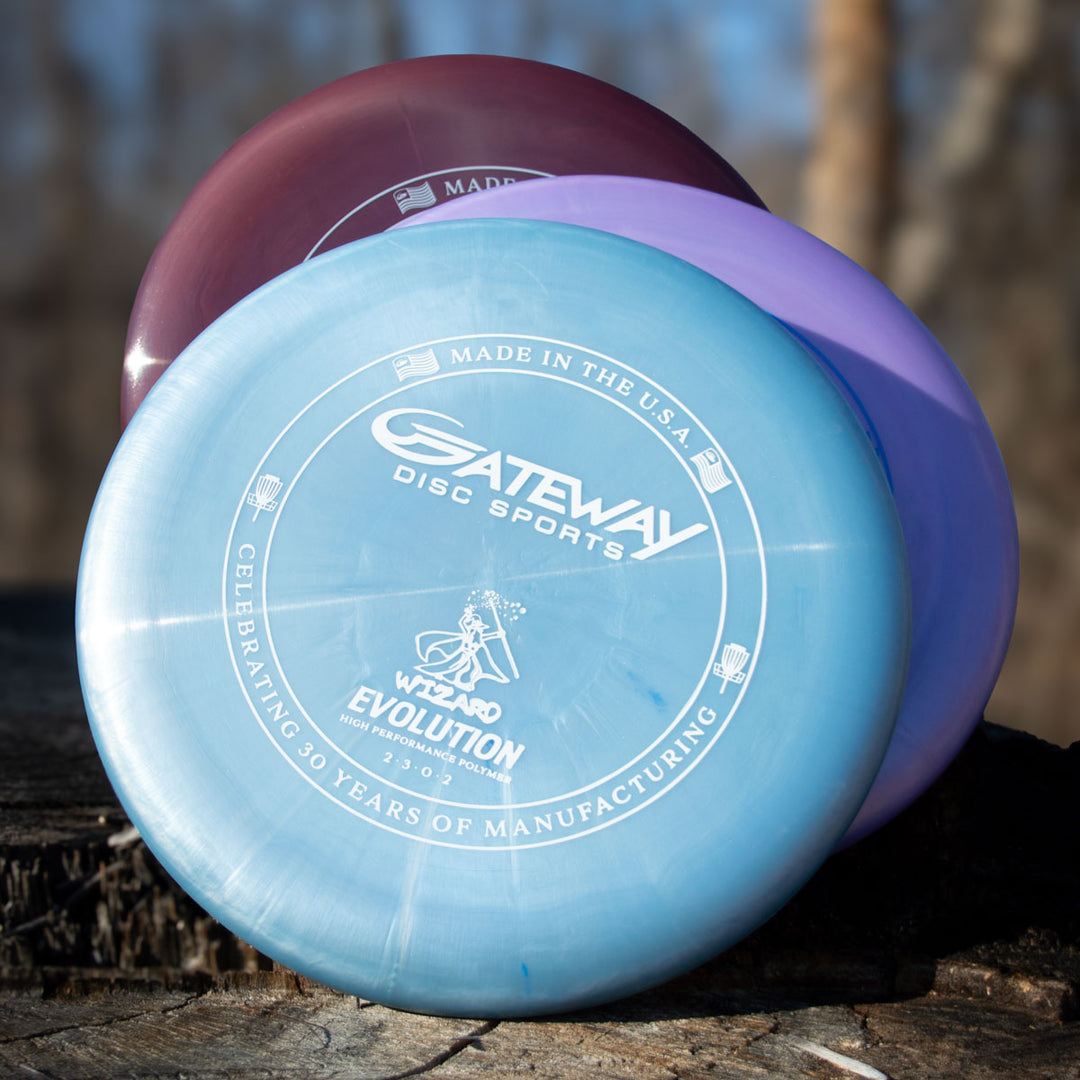 Three Wizard - Evolution - Galaxy discs from Gateway Disc Sports in blue, purple, and maroon with a pearl shimmer finish are stacked on an outdoor tree stump, displaying the 30th Anniversary logo and text on the top disc.