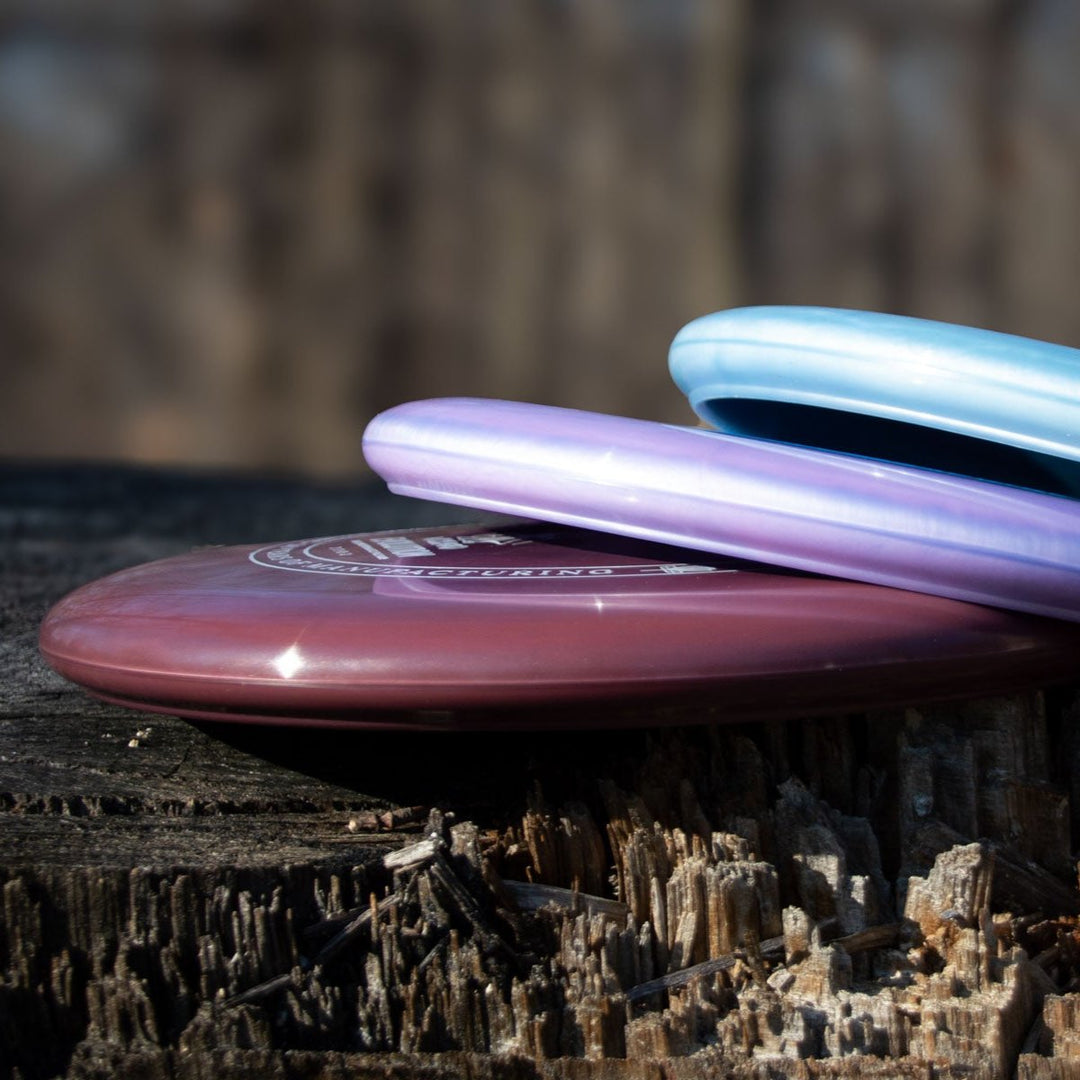 Three Wizard - Evolution - Galaxy disc golf discs—maroon, purple, and light blue with a pearl shimmer—stacked on a weathered tree stump in sunlight.