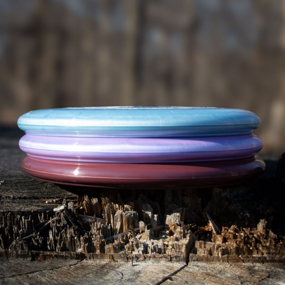 Three stacked Wizard - Evolution - Galaxy disc golf discs in blue, purple, and maroon with a pearl shimmer finish rest on rough wood outdoors, their colors popping against a blurred forest—perfect for fans of this special edition.