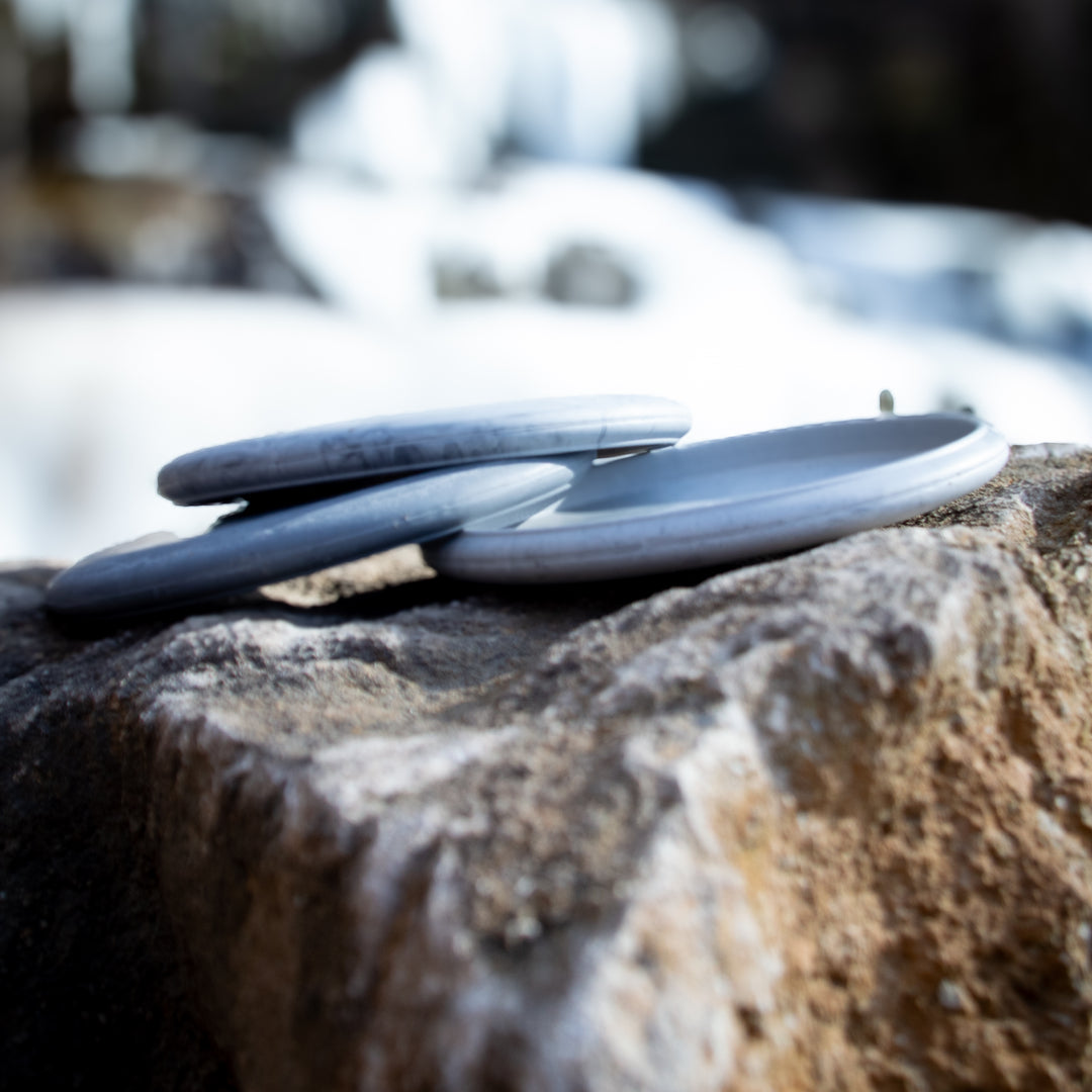 Three round, gray stones are stacked on a larger rock outdoors, their smooth look reminiscent of the Lunar blend of a Wizard putt & approach disc, against a softly blurred background.