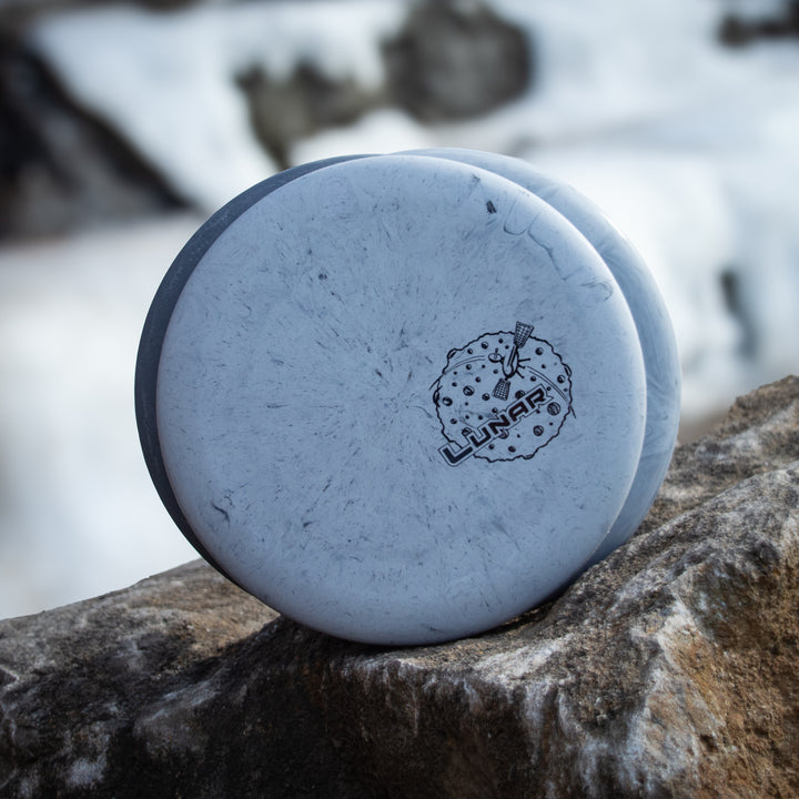 A Wizard - Lunar putt & approach disc, made from SSS plastic blend with a "Lunar" logo, rests against a rock in a snow-covered setting.