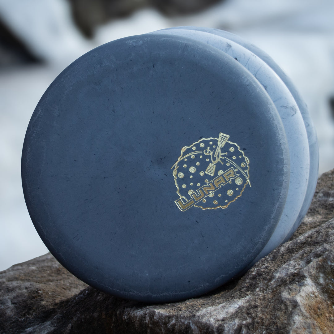 A blue-gray Wizard - Lunar putt & approach disc in SSS plastic, featuring a lunar-themed design with "LUNAR" in gold, is shown leaning against a rock outdoors.