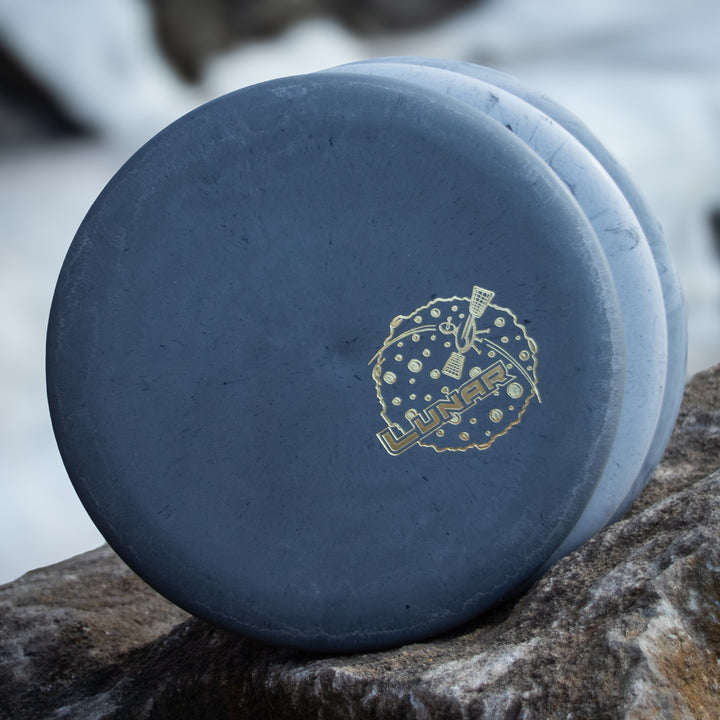 A blue-gray Wizard - Lunar putt & approach disc in SSS plastic, featuring a lunar-themed design with "LUNAR" in gold, is shown leaning against a rock outdoors.