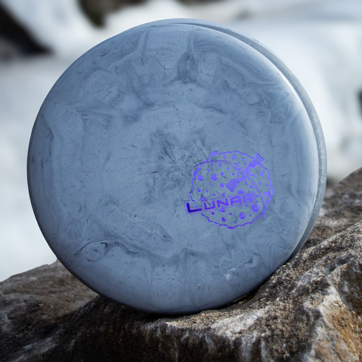 A gray Wizard - Lunar putt & approach disc in SSS plastic with a purple moon logo sits on a rock, snowy background softly blurred.