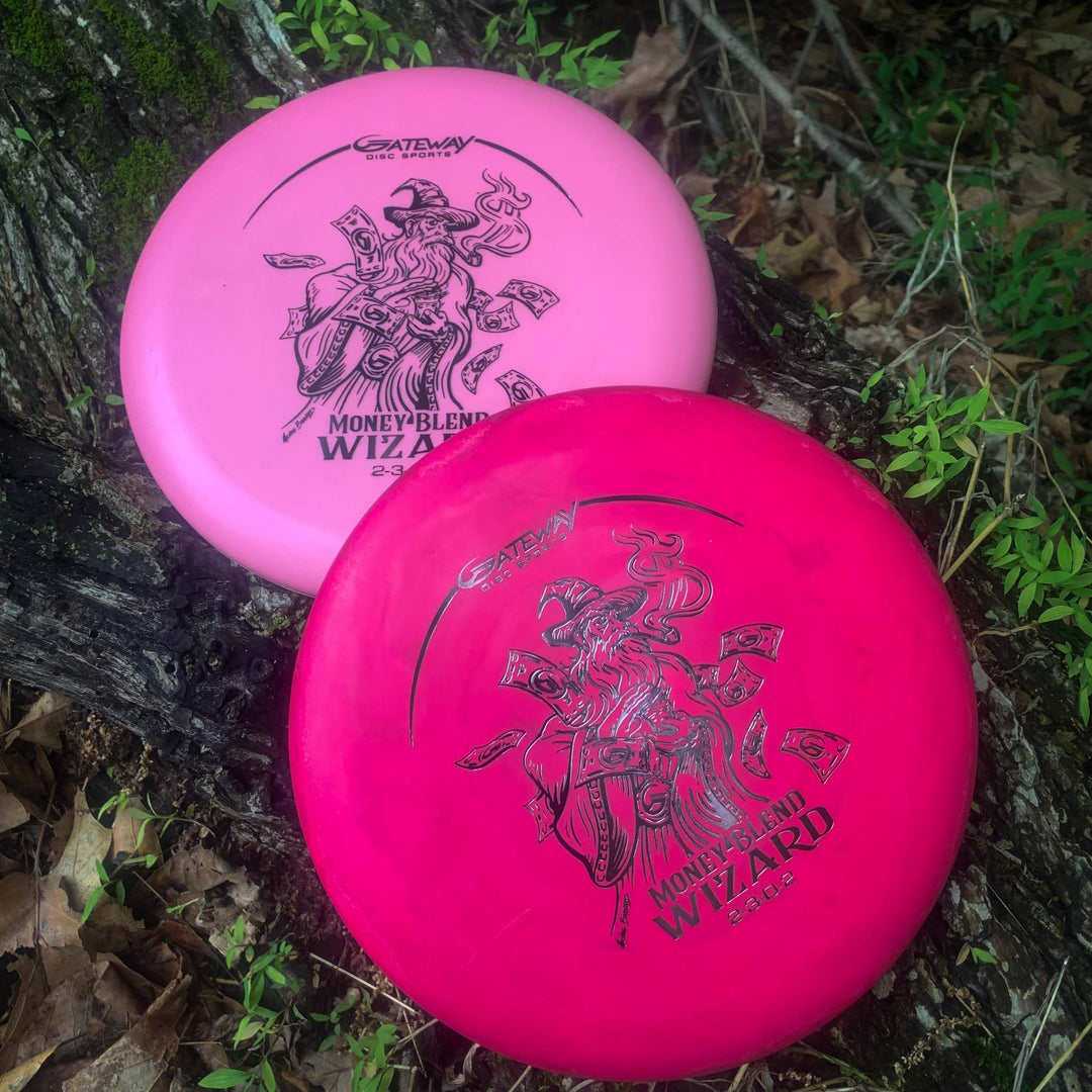 Two pink Wizard - Money putters, known for accuracy and stability, with "Money Blend" and wizard graphics, rest on the forest floor near a tree trunk.