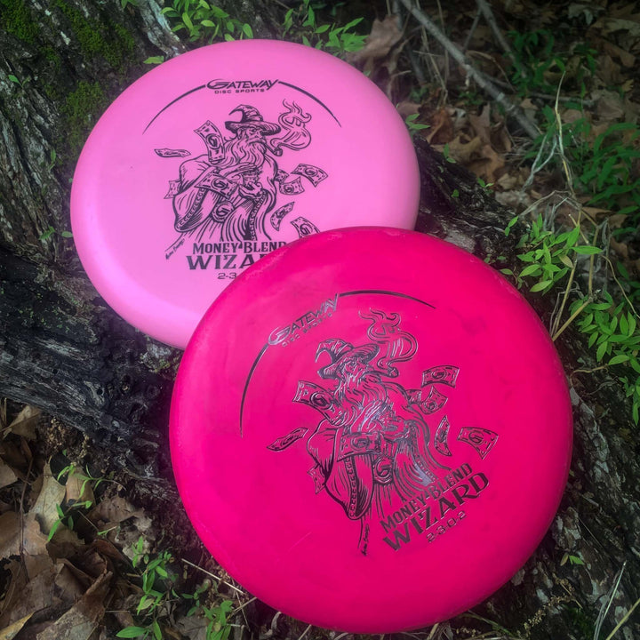 Two pink Wizard - Money putters, known for accuracy and stability, with "Money Blend" and wizard graphics, rest on the forest floor near a tree trunk.