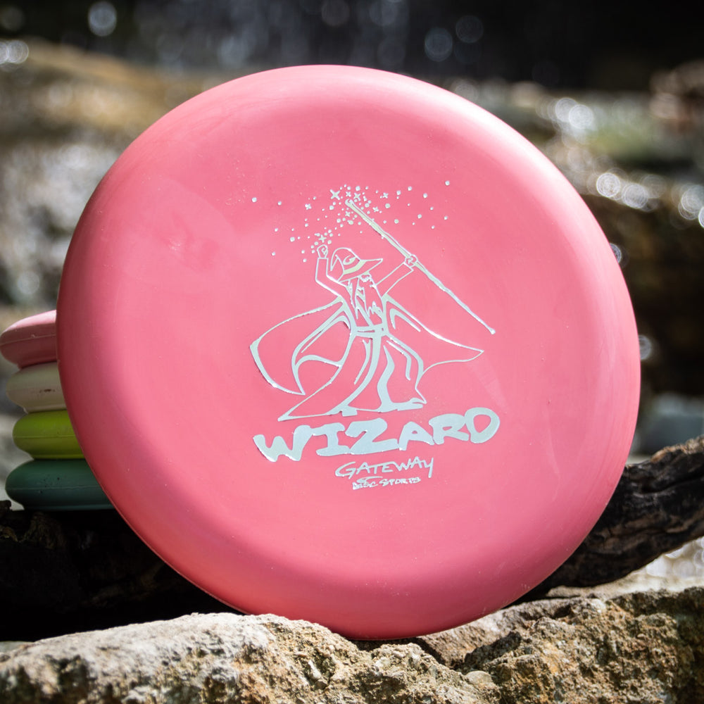 A pink Wizard - Multi Flex 5 pack putter, featuring a wizard graphic, sits on a rock outdoors by a stream.