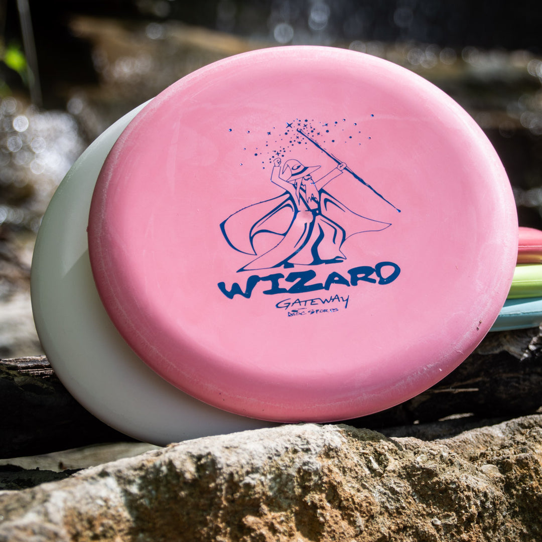A Wizard - Multi Flex 5 pack of pink disc golf putters, each featuring a wizard illustration, is displayed on a rock outdoors against a white disc. These stable putters are ideal for consistent and reliable throws.