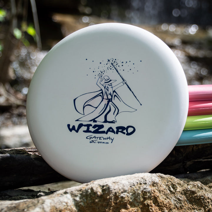 The Wizard - Multi Flex 5 pack features stable white putters with a wizard design, resting on a log outdoors beside other colored discs. Enjoy reliable performance and multi-flex 5 versatility for any disc golf game.