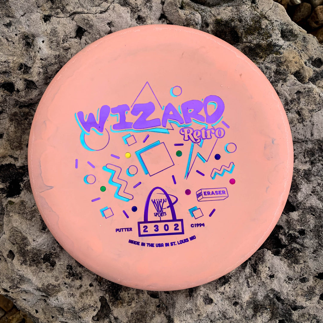 A Retro Wizard - Eraser putter with geometric designs, pink in color, is displayed on a rocky surface.