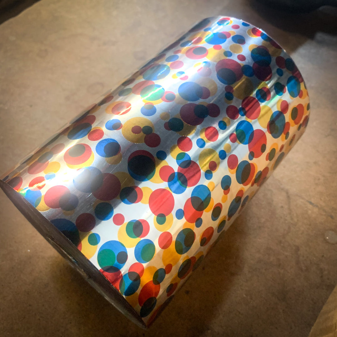 A roll of Ultra Premium Foil gift wrap, featuring a colorful polka dot pattern inspired by platinum discs, rests on a brown surface.