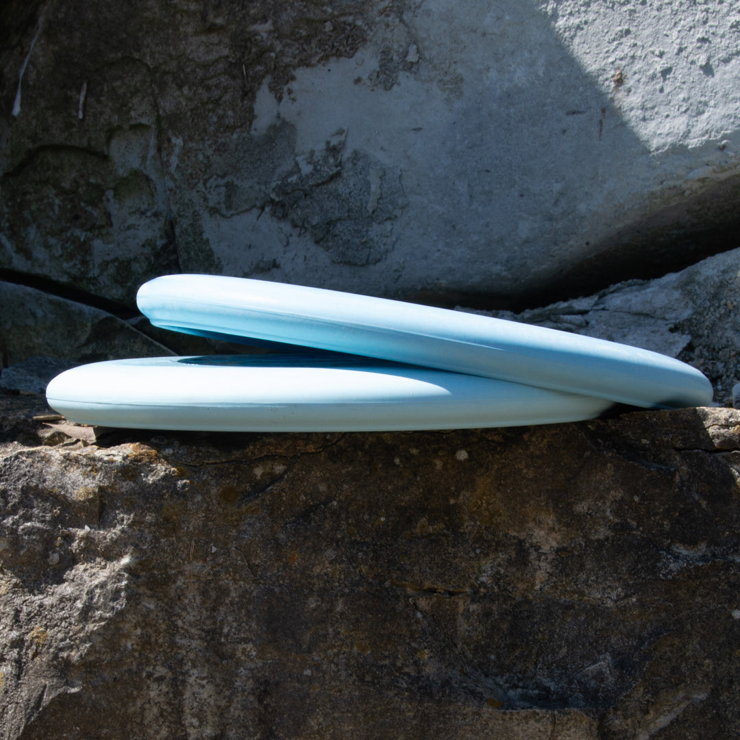 Two light blue Tyler Wozniak's "Wozard" Nylon Alloy frisbees rest on a large, rough rock surface in partial sunlight—a stable workhorse putter and excellent off the tee for mid-range drives.