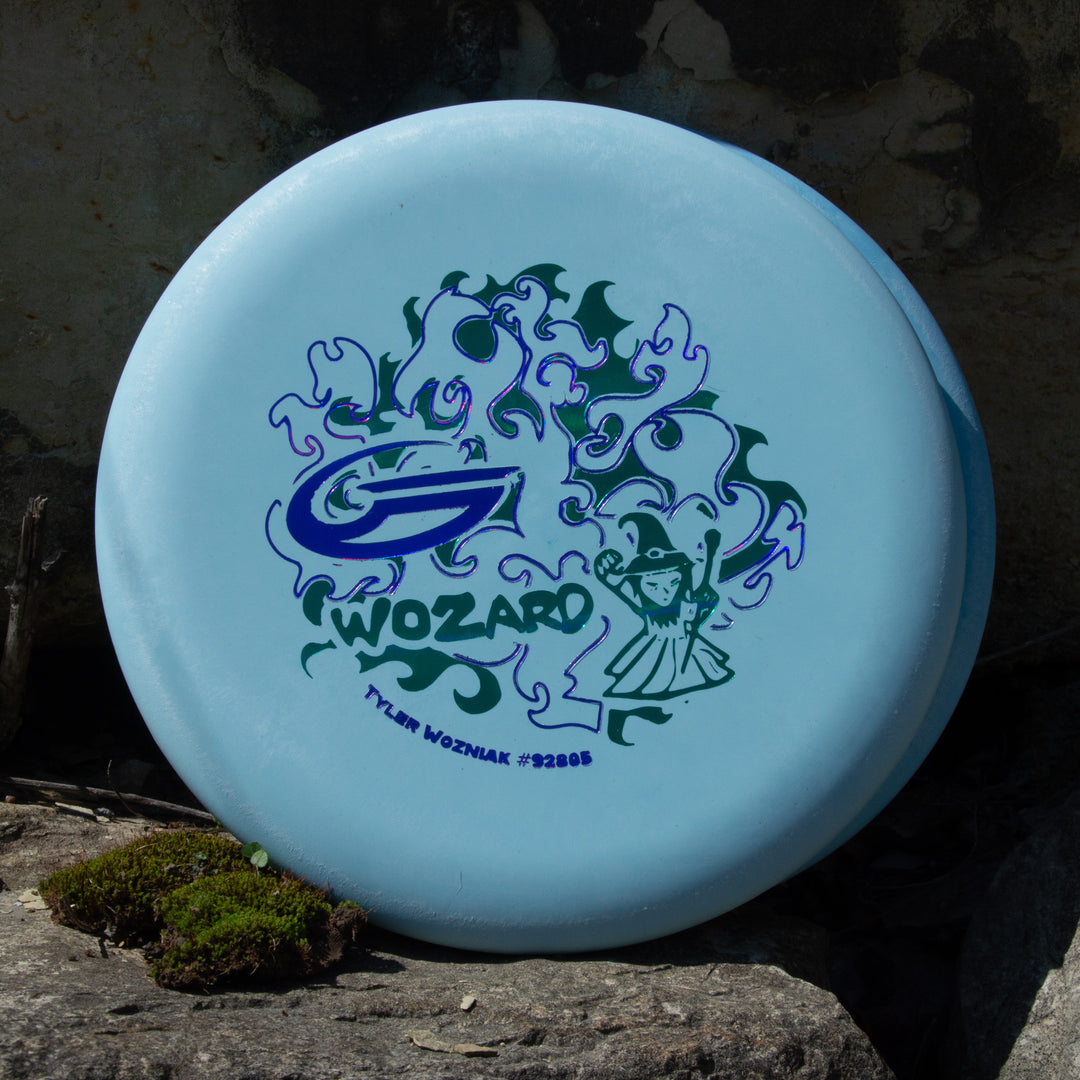 Tyler Wozniak's "Wozard" - Nylon Alloy is a light blue disc golf disc for mid-range drives, featuring flames, a wizard graphic, and "WOZARD" with "TYLER WOZNIAK #32861," displayed on a rock near moss.